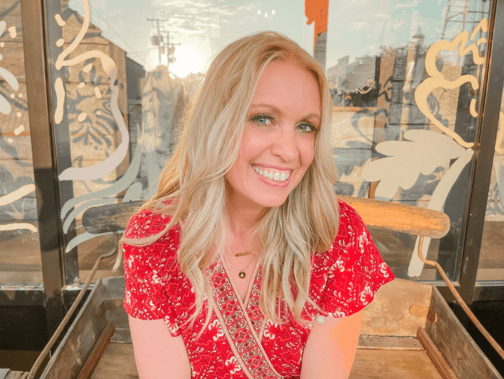 Portrait of business coach Julie Kinne smiling while sitting outdoors, wearing a red patterned wrap dress with gold necklaces, against a window background with warm sunlight.